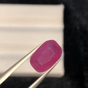 May include: A vibrant, cushion-cut ruby gemstone with a deep pinkish-red hue and faceted surfaces, held securely by a pair of silver-toned metal tweezers. The gemstone displays a translucent quality and a polished finish, showcasing its geometric shape.