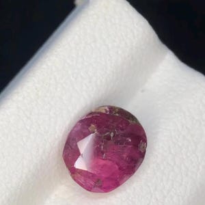 May include: A faceted oval gemstone with a deep magenta and pink hue. The stone features a polished surface with visible internal inclusions and natural variations in color, displaying a mix of translucent and opaque sections throughout its structure.