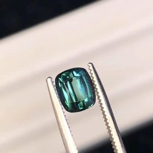 Puede incluir: Una piedra preciosa de corte cojín con una paleta de colores verde azulado y verde bosque, con una superficie facetada que refleja la luz. La piedra está sujeta firmemente por unas pinzas de metal plateado con bordes dentados en las puntas.