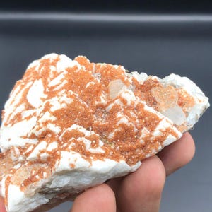 Könnte beinhalten: Ein rohes Mineralexemplar mit einem starken Kontrast zwischen undurchsichtigen weißen Kristallstrukturen und leuchtenden, texturierten orangefarbenen Ablagerungen. Der Stein zeigt eine unregelmäßige, gezackte Oberfläche mit Ansammlungen durchscheinender Formationen.