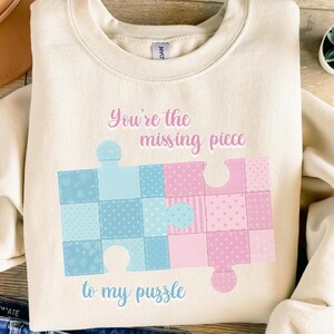 Könnte beinhalten: Cremefarbenes Sweatshirt mit einem Design aus blauen und rosa Puzzleteilen. Der Text "You're the missing piece to my puzzle" ist oberhalb und unterhalb der Puzzleteile aufgedruckt. Die Puzzleteile haben ein Patchworkmuster.