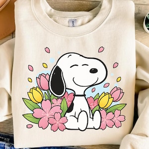 May include: Cream-colored sweatshirt featuring a smiling Snoopy character surrounded by pink and yellow flowers and green leaves. The design includes pink petals and a light blue sky background.