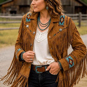 May include: A brown suede jacket featuring long fringe detailing, silver-toned metal buttons, and colorful geometric beadwork on the shoulders and cuffs. The jacket is worn over a white top with a lace neckline and a brown leather belt with a turquoise buckle.