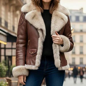May include: A brown leather jacket with a cream-colored fur collar and cuffs. The jacket has a quilted design on the shoulders and a small pocket on the chest. The model is wearing a black sweater and blue jeans.