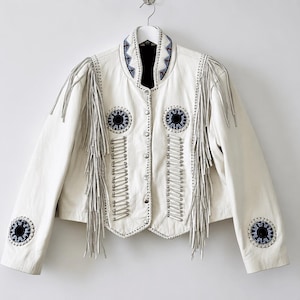 May include: A white leather jacket with silver fringe and decorative blue and black circular accents. The jacket has a round collar with blue and white patterned trim and silver metal snaps down the front. The sleeves have fringe and circular accents.