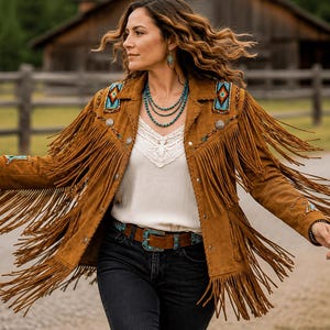 May include: A tan suede jacket featuring long fringe detailing along the sleeves and chest, accented with turquoise-colored beaded patches on the shoulders and cuffs, paired with a white lace-trimmed top, dark jeans, and a brown leather belt.