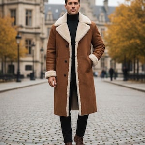 May include: A long brown coat with a cream shearling lining and trim. The coat has a double-breasted design and is worn with a black turtleneck and trousers. The person is wearing brown leather boots. The background shows a cobbled street and buildings.