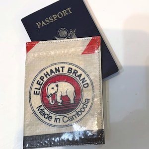 May include: A passport wallet with the text "ELEPHANT BRAND" and an elephant logo, made in Cambodia. The wallet is beige with red accents and a black border. A blue passport is partially visible behind the wallet.