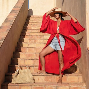 May include: A woman wearing a red kimono-style robe, denim shorts, and a straw hat stands on a terracotta staircase. A woven straw tote bag rests on the steps. The robe is tied at the waist, and the outfit is completed with sandals.