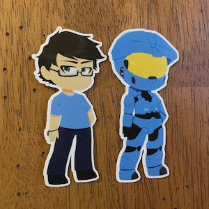 May include: Two stickers on a wooden surface. One sticker depicts a cartoon character with glasses, a blue shirt, and dark pants. The other sticker is a blue armored figure with a yellow visor. Both stickers have a white border.