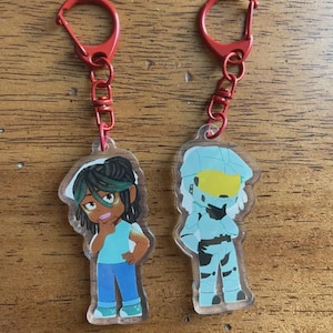 May include: Two acrylic keychains with red metal clasps. One keychain features a cartoon character with dark hair, a blue shirt, and blue jeans. The other keychain depicts a blue and yellow cartoon character in armor.