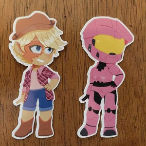 May include: Two cartoon stickers. One features a character in a brown hat, pink plaid shirt, blue shorts, and brown boots. The other is a pink armored figure with a yellow visor and black accents.