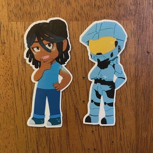 May include: Two die-cut stickers on a wooden surface. One sticker features a cartoon person in blue clothing, with dark hair and a thoughtful expression. The other sticker depicts a blue armored figure with a yellow visor, also in a thinking pose.