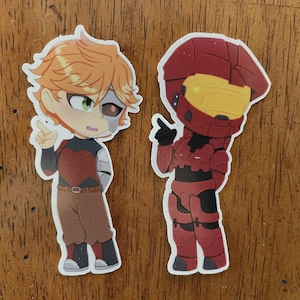 May include: Two cartoon stickers on a wooden surface. One sticker features a character with orange hair, a robotic eye, and a red and black outfit. The other sticker depicts a character in red armor with a yellow visor.