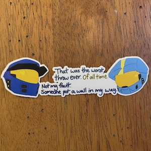 May include: A sticker featuring two cartoon helmet designs in blue and yellow, with the text "That was the worst throw ever. Of all time. Not my fault. Someone put a wall in my way."