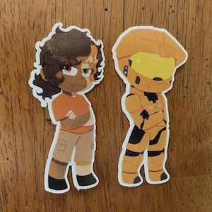 May include: Two cartoon stickers on a wooden surface. One sticker depicts a character with brown hair, an orange shirt, and khaki pants. The other sticker shows a character in orange armor with a yellow visor.