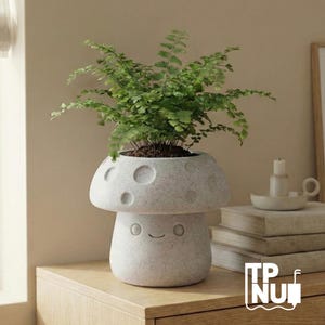 Puede incluir: Una maceta caprichosa en forma de hongo gris claro, llena de una vibrante planta verde. La maceta tiene una superficie texturizada con hendiduras circulares y un diseño de cara sonriente. La maceta está sobre una superficie de madera.
