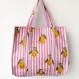 May include: A quilted tote bag featuring a pattern of vertical pink and white stripes with a repeating print of yellow lemons and green leaves. The bag includes two matching striped fabric handles and a quilted texture throughout the exterior.