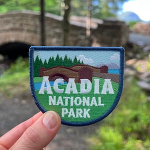 May include: A blue and green embroidered patch with the text "Acadia National Park" and a depiction of a bridge over a body of water.
