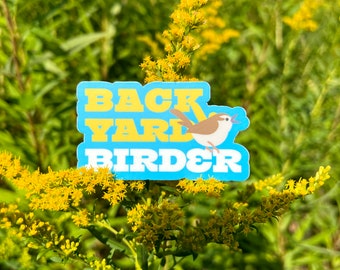 Puede incluir: Una pegatina azul y amarilla con el texto "Backyard Birder" y un pájaro de dibujos animados cantando. La pegatina está en una planta verde con flores amarillas.