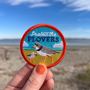 May include: A round, orange and blue embroidered patch with the text "Protect the Plovers" and an illustration of a plover bird and a smaller bird.