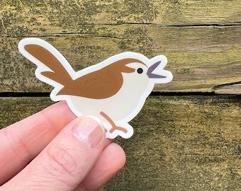 May include: A sticker featuring a cartoon bird with brown wings and head, a white belly, and a purple open beak. The bird is on a white background with a slight shadow. The sticker is held by a hand against a weathered wood background.