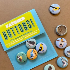 May include: A set of eight 1-inch pin-back buttons featuring colorful illustrations of birds. The buttons are packaged in a clear plastic bag with a blue and yellow label that reads "FATBIRD BUTTONS! One-inch pin back button set FATBIRDDESIGN.COM MADE IN THE USA".