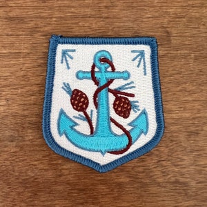 May include: An embroidered blue anchor with a brown rope and pine cones on a white background. The anchor is surrounded by a blue border and is in the shape of a shield.