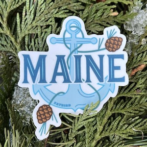 以下が含まれることがあります： ロープのデザインが施された青い錨と、青い文字で「MAINE」と書かれています。錨の両側に2つの茶色の松ぼっくりがあります。錨は白い背景の上にあります。