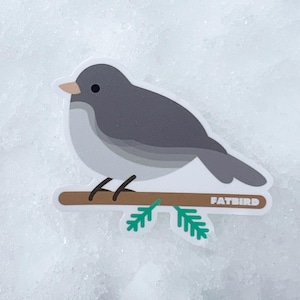 May include: A gray bird with a brown beak perched on a brown branch with green leaves. The bird is sitting on a white background. The sticker says "Fatbird" on the branch.