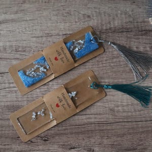 Może przedstawiać: Dwie ręcznie robione zakładki do książek z niebieskimi i srebrnymi akcentami, każda z frędzlem. Zakładki są zapakowane w brązowy papier z napisem "Handmade with love" i czerwonym sercem. Tło to drewniana powierzchnia.