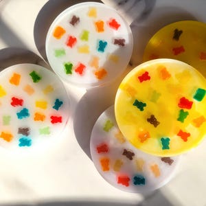 Może przedstawiać: Kolekcja kolorowych podkładek z żywicy. Okrągłe podkładki zdobione są kształtami żelków w kolorach czerwonym, pomarańczowym, żółtym, zielonym i brązowym. Niektóre podkładki są białe, inne żółte.