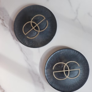 Może przedstawiać: Dwa ciemnoszare podkładki pod szklanki ze złotym, przeplatającym się wzorem. Podkładki są okrągłe i mają metaliczny połysk. Leżą na białej marmurowej powierzchni.