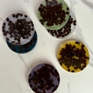 Może przedstawiać: Zestaw okrągłych podkładek z żywicy, z zatopionymi ziarnami kawy. Podkładki są w różnych kolorach, takich jak niebieski, zielony, żółty i fioletowy. Ziarna kawy mają ciemnobrązowy kolor, tworząc kontrast.