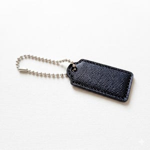 May include: A navy blue leather tag with a textured finish, connected to a silver ball chain. The tag is rectangular with rounded corners and a small hole for the chain. The background is white.
