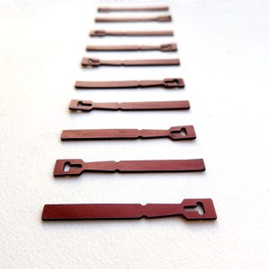 May include: A series of ten flat, reddish-brown strips arranged in a vertical line, each featuring a T-shaped cutout at one end and a small notch along the side. These uniform, elongated components are laid out in a sequence from smallest to largest.