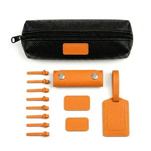 May include: A black zippered pencil case with a textured surface and an orange rectangular patch, accompanied by eight orange cable ties, a small orange snap-button wrap, two small orange rectangular tags, and one orange luggage tag.