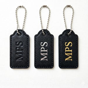 May include: Three black luggage tags with silver ball chain attachments. Each tag has the initials "MPS" in a different color: embossed, silver, and gold. The tags have a textured surface.