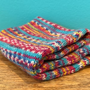 May include: Folded knitted textile in shades of teal, pink, yellow, and blue. The fabric has a textured appearance with a repeating pattern of small stitches. The item is stacked on a wooden surface against a teal background.