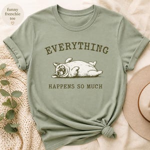 May include: Sage green t-shirt featuring a relaxed bulldog illustration with the text "EVERYTHING HAPPENS SO MUCH". The shirt has rolled-up sleeves and a knotted hem. A beige hat and a cream-colored knitted blanket are in the background.