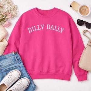 May include: A vibrant pink sweatshirt with the text "DILLY DALLY" in white, paired with blue jeans and white sneakers. Accessories include sunglasses, a beige purse, and a smartphone.