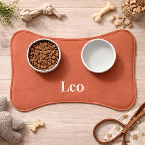 Peut inclure: Tapis de nourriture pour animaux de compagnie orange en forme d'os avec le nom "Leo" en blanc. Le tapis contient deux bols blancs, l'un rempli de croquettes brunes et l'autre vide. Un jouet pour chien, des friandises et une laisse sont &eacute;galement visibles.