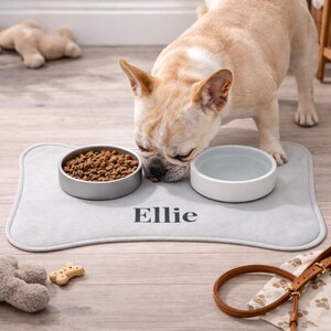 Peut inclure: Un tapis de nourriture pour chien en forme d'os gris clair avec le nom "Ellie" imprim&eacute; en noir. Le tapis contient deux bols, l'un avec de la nourriture pour chien et l'autre avec de l'eau. Un bouledogue fran&ccedil;ais mange dans le bol de nourriture.