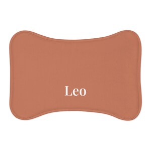 Peut inclure: Un lit pour animaux de compagnie de couleur terre cuite en forme d'os. Le mot "Leo" est imprim&eacute; en blanc au centre du lit. Le lit a des bords arrondis et une apparence douce et moelleuse.