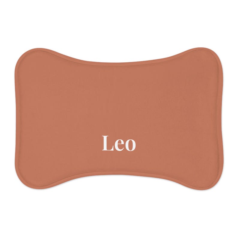 Peut inclure: Un lit pour animaux de compagnie de couleur terre cuite en forme d'os. Le mot "Leo" est imprim&eacute; en blanc au centre du lit. Le lit a des bords arrondis et une apparence douce et moelleuse.