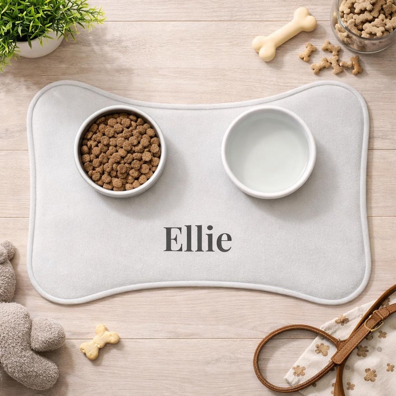 Peut inclure: Tapis de gamelle pour animaux de compagnie en forme d'os, gris clair, avec deux bols blancs, l'un rempli de nourriture pour chiens et l'autre d'eau. Le nom "Ellie" est imprim&eacute; sur le tapis. Accessoires : un os et une laisse.