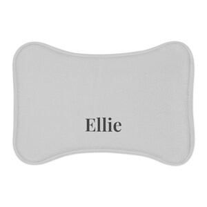 Peut inclure: Un lit pour animaux de compagnie en forme d'os, gris clair, avec le nom "Ellie" imprim&eacute; en noir. Le lit a un bord doux et arrondi. Le design est simple et moderne.