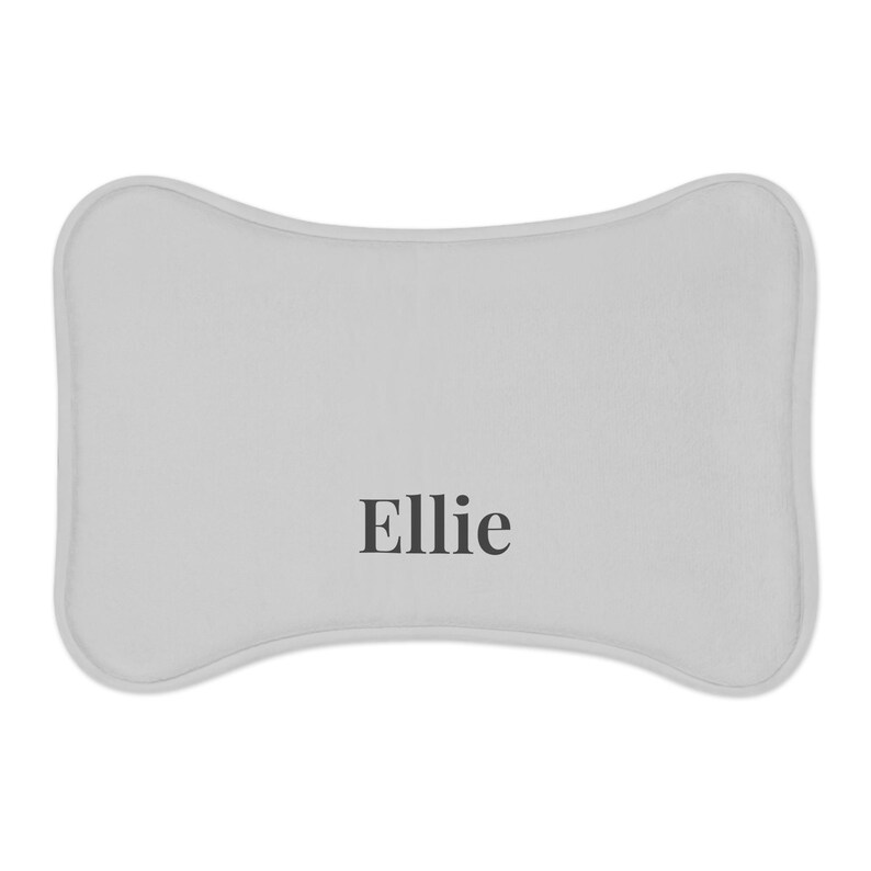 Peut inclure: Un lit pour animaux de compagnie en forme d'os, gris clair, avec le nom "Ellie" imprim&eacute; en noir. Le lit a un bord doux et arrondi. Le design est simple et moderne.