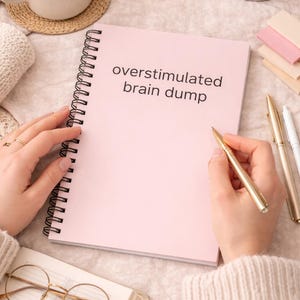 Könnte beinhalten: Ein rosa Spiralnotizbuch mit den Worten "overstimulated brain dump" auf dem Cover. Ein goldener Stift wird über dem Notizbuch gehalten. Weitere Artikel sind eine Tasse Kaffee, eine Kerze und eine Brille.