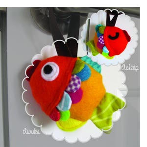 May include: A colourful, plush fish toy with a red mouth and a large eye. The fish has a removable, red belly that can be flipped to reveal a different side with colourful spots. The toy is labelled "awake" and "asleep" on the sides.